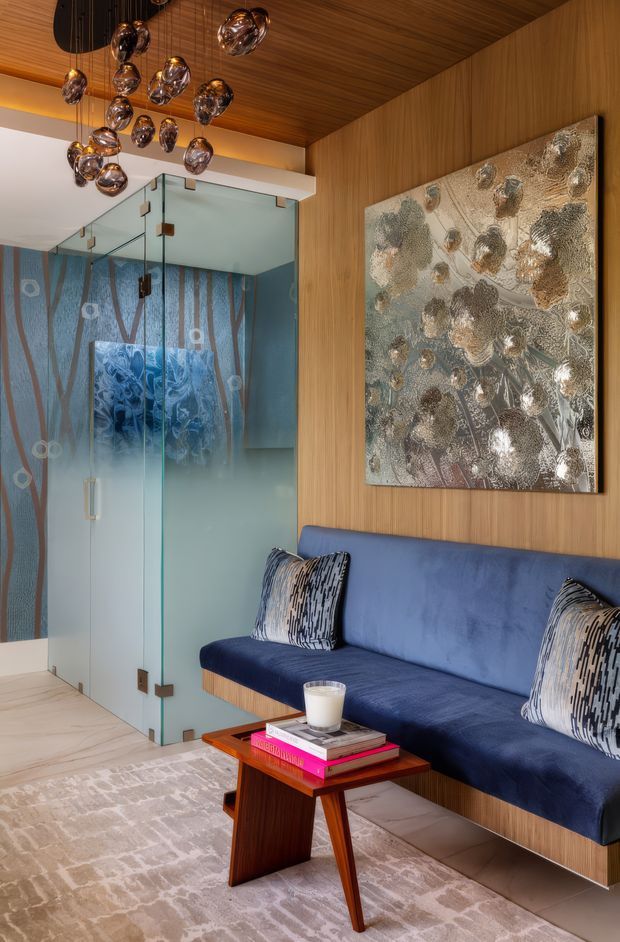 A refined contemporary living room or lounge with a wall-mounted deep blue upholstered sofa set against warm vertical timber wall panelling. A sculptural glass-and-metal pendant cluster hangs from the timber ceiling, while a textured metallic abstract artwork provides a feature wall accent. Frosted glass doors with decorative blue patterned glazing open toward an adjacent space. A small angular timber coffee table, books, cushions, and a pale patterned rug complete the sophisticated interior.