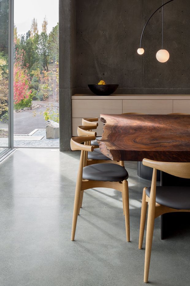 A contemporary dining area with a large live-edge timber dining table and sculptural wooden dining chairs with dark upholstered seats. The space features a polished concrete floor, a dark exposed concrete feature wall, pale built-in cabinetry, and two minimalist globe pendant lights. Full-height glazing opens to a landscaped garden outside, bringing in abundant natural light.