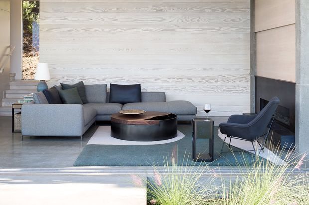 A refined contemporary living room with a grey sectional sofa, dark sculptural round coffee table, lounge chair, side table, and floor lamp. The room features a pale horizontal timber feature wall, polished concrete flooring, a large fireplace with dark surround, and expansive glazing opening toward planted landscaping. The palette is predominantly soft grey, pale timber, charcoal, and muted blue-green.