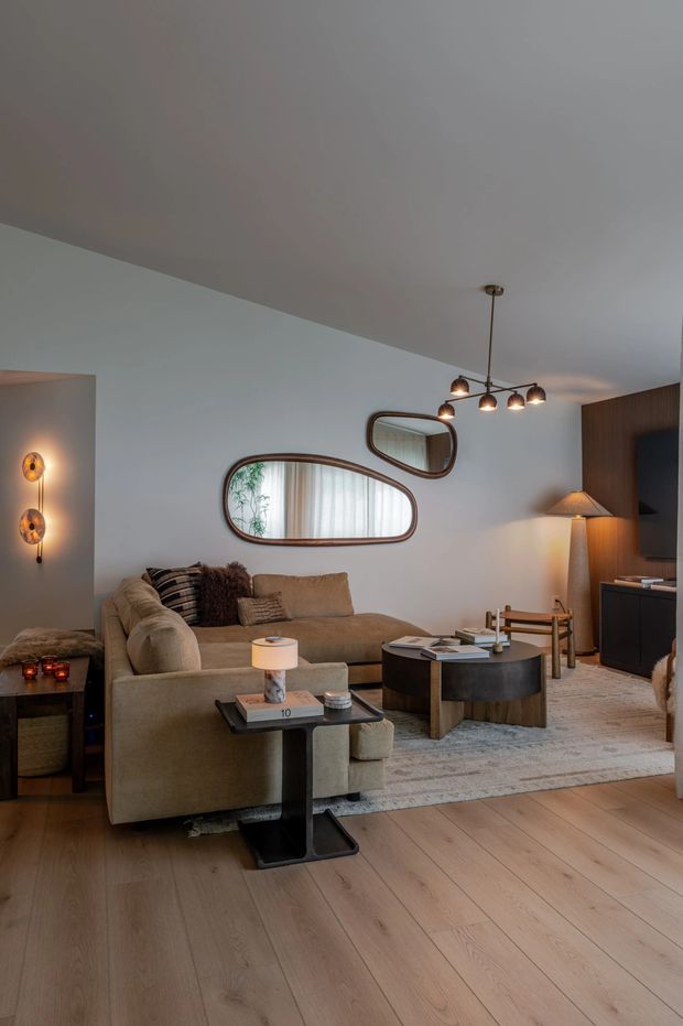 A warm, refined living room with a large curved sectional sofa, layered neutral textiles, round dark coffee table, sculptural side tables, and a textured area rug. Pale timber floorboards contrast with white walls and a dark timber media wall. Organic-shaped framed mirrors, a linear pendant chandelier, wall sconces, and table lamps provide soft ambient lighting.