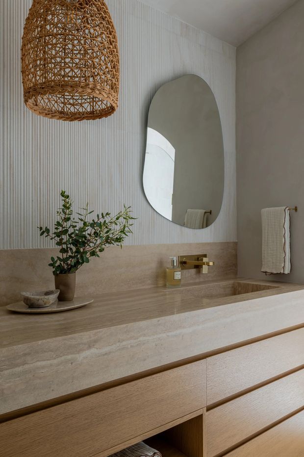 A refined bathroom vanity area featuring a large integrated stone benchtop and backsplash, warm timber cabinetry, a wall-mounted brass tap, an organic-shaped mirror, and a woven pendant light. The wall is finished with vertical white textured panelling, complemented by neutral stone tones and understated natural styling.