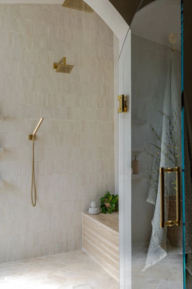 A refined walk-in shower with warm cream textured wall tiles and matching stone-look floor tiles. Brushed brass shower fittings, handheld shower, glass enclosure, and a built-in stone bench with a small plant create a calm, spa-like bathroom interior.