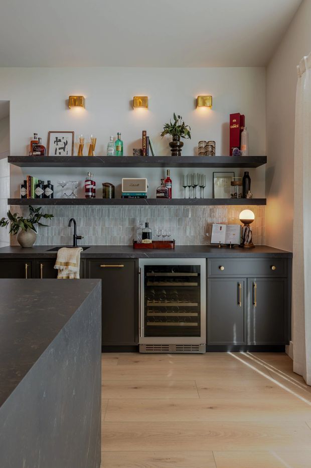 A sophisticated home bar and kitchenette with dark charcoal built-in cabinetry, a stone island, black benchtop, pale timber flooring, and a pale vertically textured tile splashback. Open shelves display bottles and glassware, with three warm wall sconces above. The space includes a sink, underbench wine refrigerator, brass hardware, and layered decorative lighting, creating a refined, moody entertaining area.