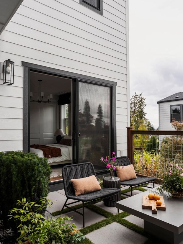 A compact contemporary outdoor terrace beside a white horizontal weatherboard-clad home. The space features black-framed sliding glass doors opening to a visible bedroom, black woven lounge chairs with neutral cushions, a low rectangular outdoor coffee table, potted flowers, clipped greenery, and a dark timber-and-wire balustrade overlooking neighbouring homes. The terrace uses large square concrete pavers separated by planted strips and has an overcast, muted atmosphere.