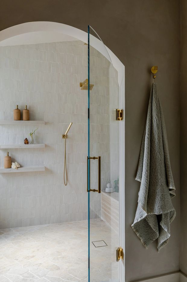 A refined bathroom shower enclosure with a curved arched opening, white textured wall tiles, pale stone floor tiles, integrated open shelving, and brass shower hardware. A frameless glass shower door with brass hinges and handle stands beside a taupe painted wall with a hanging textured towel. The overall design is warm, understated, and spa-like.