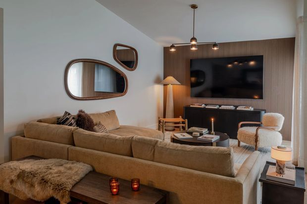 A warm, stylish living room centred on a large beige sectional sofa facing a wall-mounted television. The TV wall features dark vertical timber slats and a low dark media console, while the room includes sculptural rounded wood-framed mirrors, a floor lamp, pendant lighting, an upholstered armchair, and a dark wood coffee table. The palette is earthy and neutral with soft layered lighting and tactile textiles.