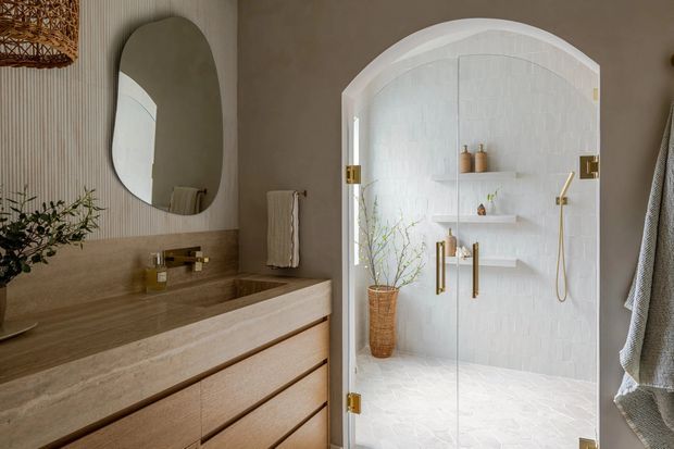 Warm, refined bathroom interior featuring a floating timber vanity with a stone benchtop, integrated basin and wall-mounted brass tapware. A rounded organic-shaped mirror hangs above the vanity, while an arched, glass-enclosed shower occupies the right side with white tiled walls, built-in shelving, brass fixtures and a woven basket. Neutral taupe walls, natural timber, and soft organic accessories create a calm contemporary feel.