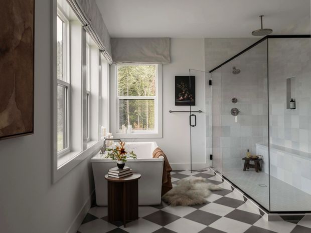 A bright, refined bathroom with a freestanding bathtub positioned beside tall windows, a large frameless glass walk-in shower with dark metal framing, and a dramatic black-and-white checkerboard floor. The room has pale grey walls and tiled shower surfaces, contemporary black fixtures, a small timber side table, and a restrained decorative scheme. A wooded landscape is visible outside the windows.