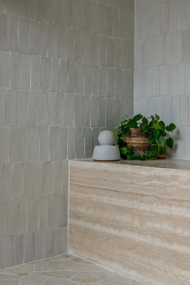 Close-up of a calm, minimalist bathroom corner featuring vertically arranged pale grey handmade-look wall tiles, a beige travertine-style tiled vanity or ledge, and a small green plant in a woven basket beside a rounded ceramic accessory. The floor uses irregular pale stone tiles with light grout.