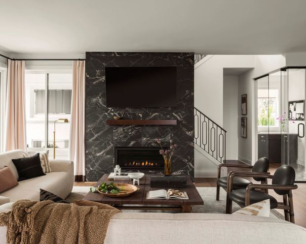 A refined open-plan living room viewed from behind a sofa, featuring a large dark marble fireplace and TV feature wall, a slim floating timber shelf, neutral upholstered seating, a timber coffee table, and a pair of sculptural leather lounge chairs. A glazed stairway and adjacent kitchen or circulation area are visible to the right, while tall windows with sheer curtains bring in daylight from the left. The space combines contemporary detailing with classic, warm furnishings.