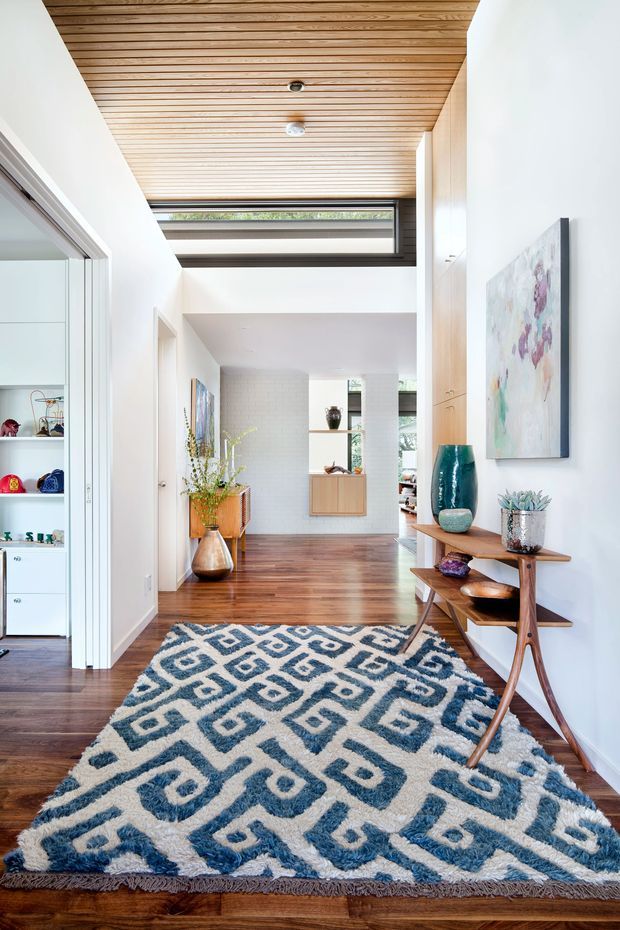 Bright residential hallway and entry passage with polished dark timber flooring, a slatted timber ceiling, white walls, high clerestory glazing, and a patterned blue-and-cream rug. A slim timber console displays decorative objects and artwork, while open shelving and adjoining rooms are visible to the side and beyond.