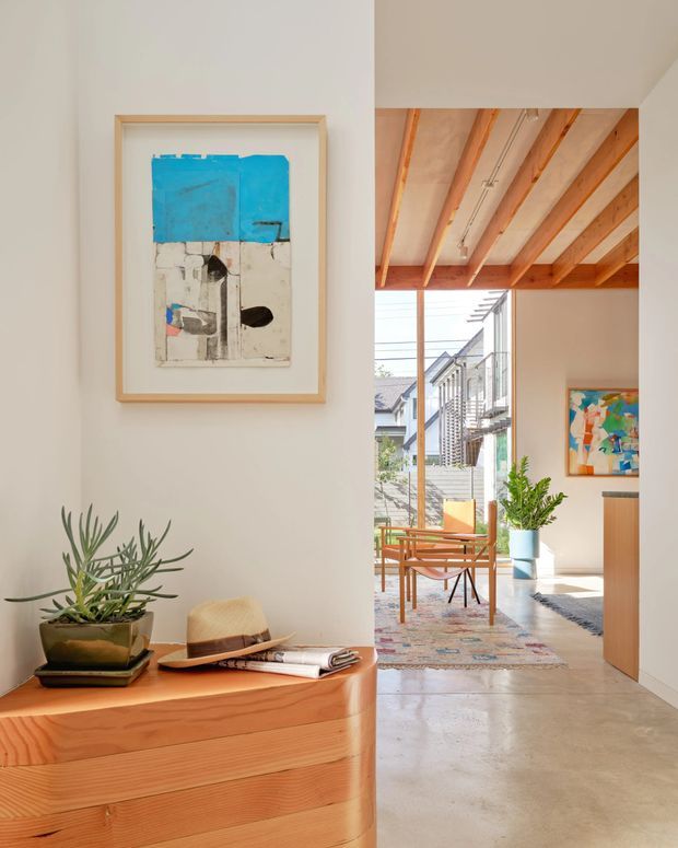 Bright contemporary interior view looking from a hallway or entry area into an open living space. A curved-edged timber console with a potted plant, hat and framed abstract artwork occupies the foreground. The adjoining room has polished concrete flooring, exposed timber ceiling beams and a large glazed opening, with timber lounge furniture, a patterned rug and additional artwork. The palette is warm, neutral and sunlit.