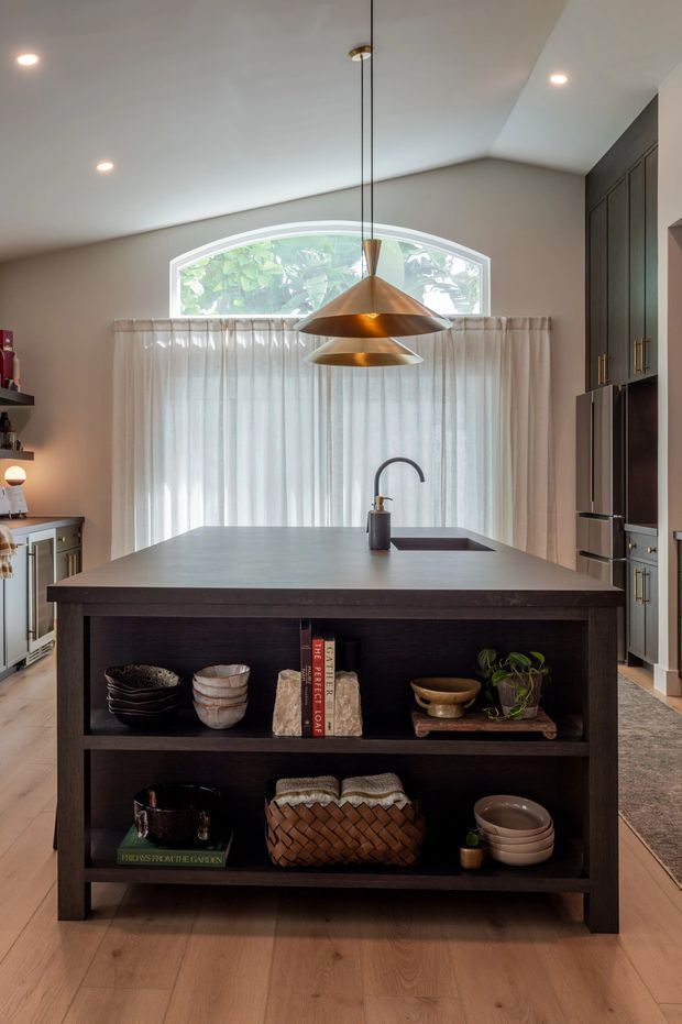 A warm, contemporary kitchen centred on a large dark timber island with open display shelving, a stone-look benchtop, undermount sink and curved black tap. Tall dark cabinetry with integrated appliances lines the right wall, while a large arched window with sheer curtains fills the rear wall. Brass pendant lights hang above the island, and pale timber flooring adds warmth.