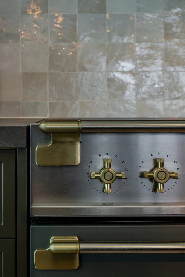Close-up detail of a premium range cooker or oven set into dark cabinetry. The appliance has brushed stainless-steel panels, prominent aged brass or bronze handles and controls, and a glossy grey-green handmade-look tiled backsplash with reflective variation.