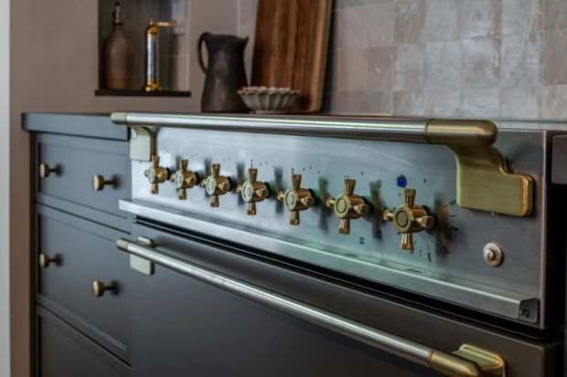 Close-up of a premium range cooker integrated into dark cabinetry, featuring a brushed stainless-steel control panel, numerous brass and gold-toned knobs, and substantial horizontal handles. A tiled wall and open shelf with decorative vessels are visible behind it.