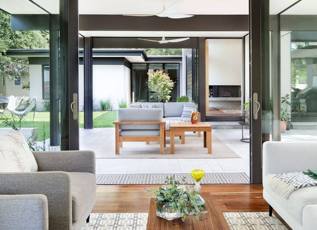 Bright contemporary living room opening through large glazed doors to a landscaped courtyard. The room features dark timber flooring, pale upholstered seating, a timber coffee table, patterned rugs, exposed dark structural beams, and strong indoor–outdoor visual connection. A secondary seating area and fireplace are visible beyond the courtyard.