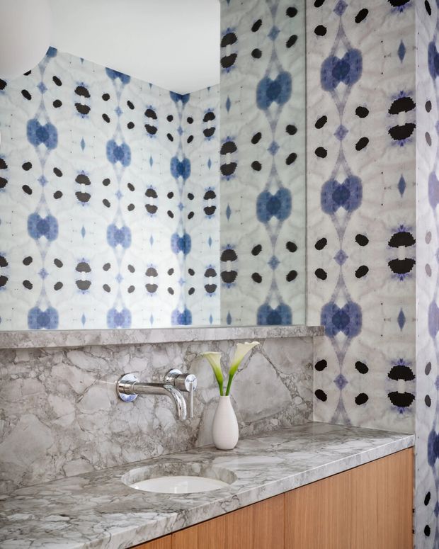Close-up view of a stylish bathroom vanity with a grey-white veined marble countertop and integrated basin, brushed metal wall-mounted tapware, and warm timber cabinetry. The walls are covered in a bold blue, black, and white patterned tile or printed surface, creating a graphic feature backdrop. A small white vase with green leaves sits on the counter.