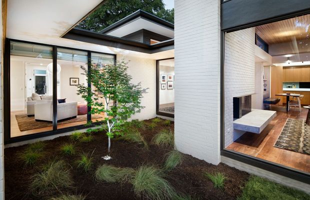 A contemporary courtyard view of a low-profile home with white-painted brick walls, broad black-framed glazing, and a sheltered planted garden. Through the windows, a living area with pale seating is visible on the left, while the right-hand interior includes a fireplace, timber flooring, a kitchen with wood cabinetry and island, and a timber-lined ceiling with pendant lighting. The composition emphasizes indoor–outdoor connection around the central courtyard.