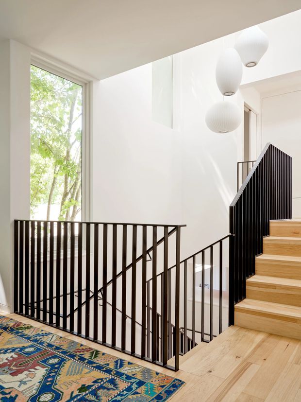 Bright contemporary stairway and landing with pale timber stairs and flooring, crisp white walls, and a slim black metal balustrade. Large glazing brings in leafy outdoor views, while three sculptural white pendant lights hang over the stair void. A colourful patterned rug adds contrast to the otherwise restrained palette.