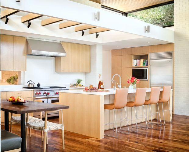 Bright, spacious contemporary kitchen with a large central island and integrated breakfast bar, pale timber cabinetry, white benchtops, stainless-steel appliances, and warm polished timber flooring. A dining table with woven chairs occupies the foreground. Exposed timber ceiling battens, clerestory glazing, and a high open ceiling add architectural interest and abundant natural light.