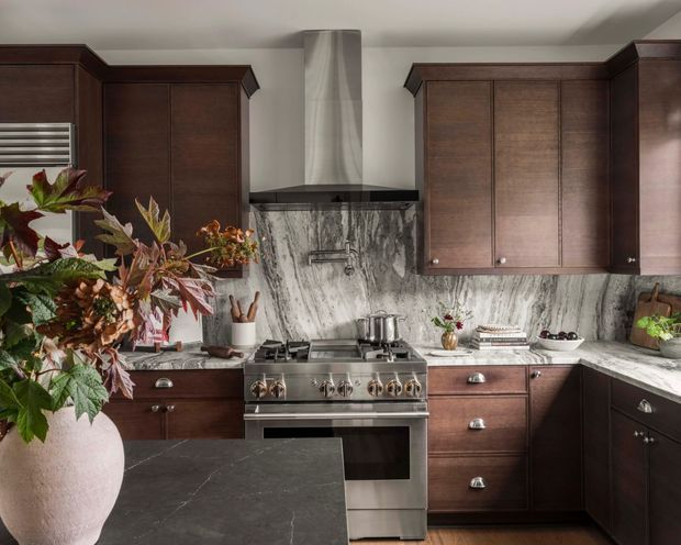A richly detailed traditional kitchen with dark stained timber cabinetry, ornate upper cabinet crown trim, a stainless-steel range and hood, and dramatic veined grey-white stone continuing across the backsplash and benchtops. A dark stone island occupies the foreground, while plants, copper-toned cookware, and decorative objects add warmth and texture.