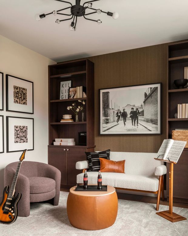A refined living room styled as a music-oriented lounge, featuring a cream sofa, rounded mauve armchair, orange leather cushion, dark timber built-in shelving and cabinetry, textured brown wall panelling, framed monochrome artwork, a guitar, sheet-music stand, and a round sculptural timber coffee table. A contemporary multi-arm ceiling light completes the warm, curated interior.