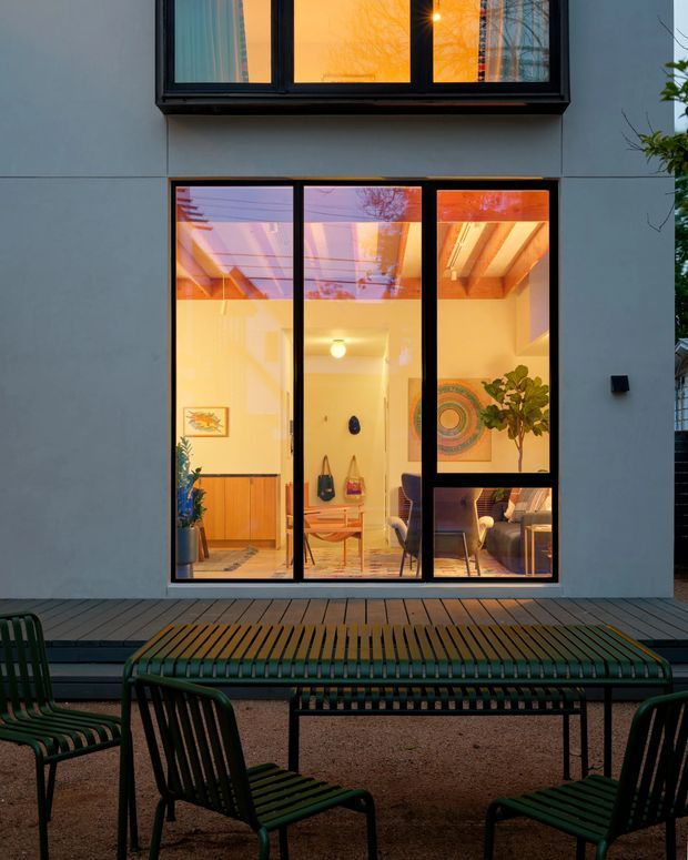 Dusk view from an outdoor terrace toward a contemporary home's tall black-framed windows and glazed doors. The illuminated interior reveals a dining area, timber ceiling beams, cabinetry, artwork and plants, while the foreground has a timber deck and dark green slatted outdoor dining furniture. The facade is smooth grey and strongly framed by large vertical glazing.