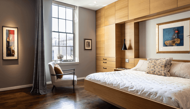 A refined bedroom with warm timber built-in cabinetry wrapping around the bed, integrated drawers and open shelving, and a low timber bed base. The room has dark painted walls, a tall divided-pane window with full-length curtains, dark timber flooring, framed artwork, an upholstered armchair, and layered neutral bedding. The overall look is warm, tailored, and contemporary with traditional architectural proportions.