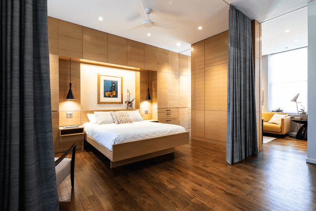 A warm contemporary bedroom with a floating timber platform bed, extensive built-in timber cabinetry and a softly illuminated recessed headboard wall. Dark full-height curtains frame the bedroom and partially separate it from a bright adjoining sitting area with a yellow armchair. The room features rich timber flooring, pendant bedside lights, recessed ceiling lights and a ceiling fan.