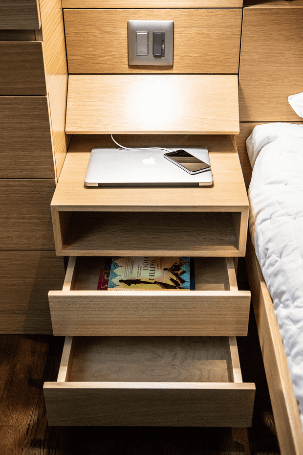 Close-up view of a built-in timber bedside table beside a bed. The unit features a light wood finish, open recessed storage, two deep drawers, a raised shelf with an electrical outlet and switches, and a fold-down charging shelf holding a laptop and phone. The bed with white bedding is visible at right, while darker timber cabinetry and flooring surround the furniture.