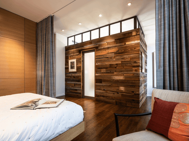 A warm, contemporary bedroom featuring a bed in the foreground, dark timber flooring, full-height curtains, clerestory glazing, and a freestanding partition or enclosed storage volume clad in reclaimed-style horizontal timber boards. The room combines white walls and bedding with rich brown wood and deep red accent cushions.