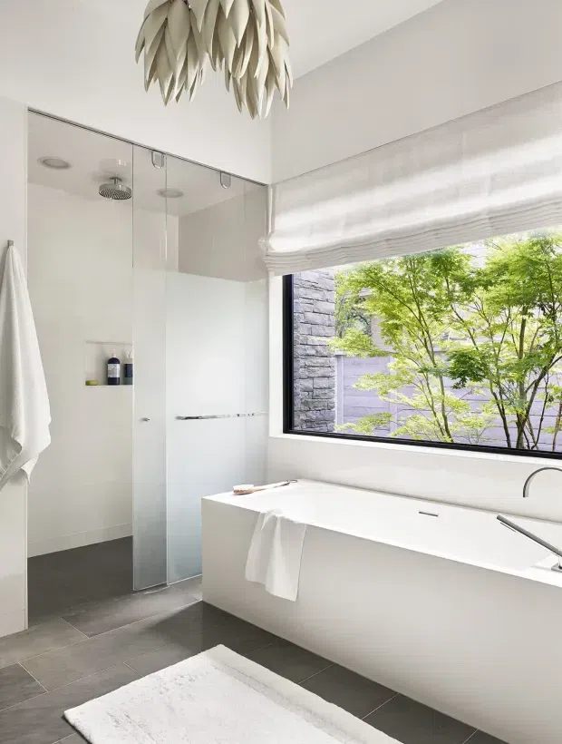 Bright contemporary bathroom featuring a freestanding white bathtub beneath a large black-framed window, a frameless glass shower enclosure, dark grey floor tiles, white wall finishes, and a sculptural leaf-like pendant light. The window overlooks a landscaped courtyard with a stone wall and tree.