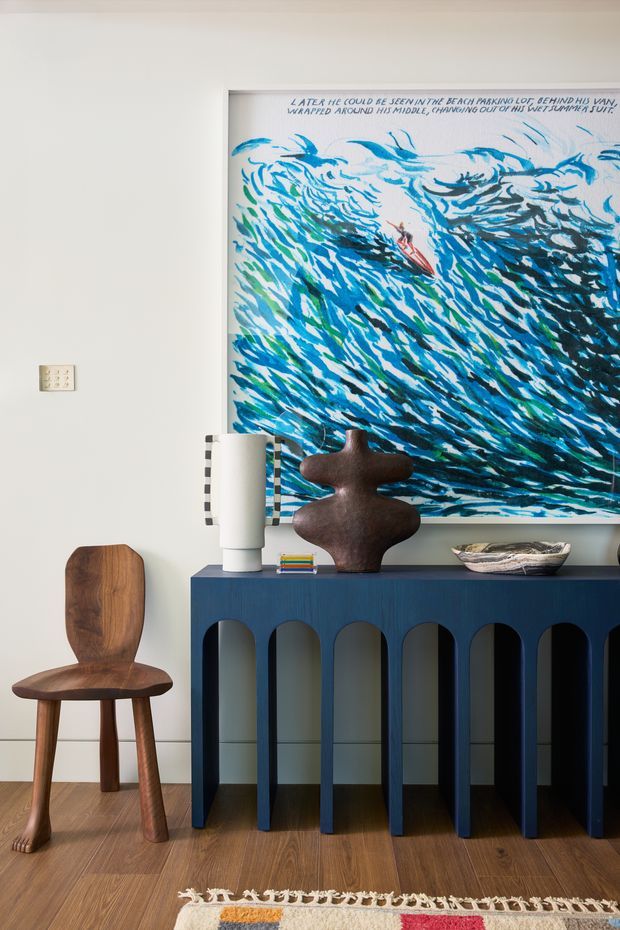 A styled interior vignette featuring a sculptural dark-blue console with repeated arched openings, a large expressive blue-and-teal ocean artwork, a carved wooden chair, sculptural brown decor, a white table lamp, and a shallow ceramic bowl. The room has pale walls, warm timber flooring, and a small patterned rug, creating an eclectic, art-focused atmosphere.