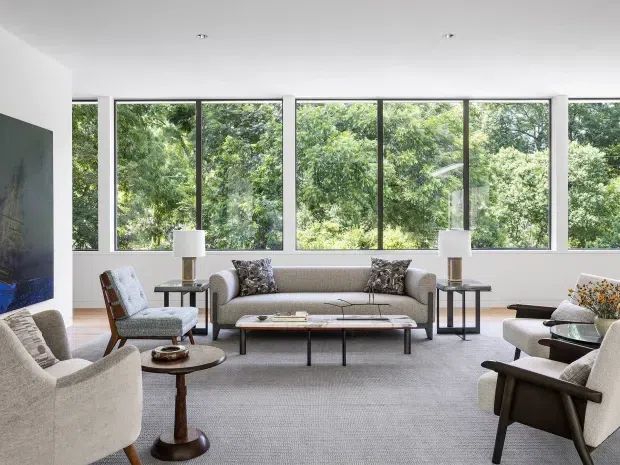Bright contemporary living room with a low-profile upholstered sofa and armchairs arranged around a slim rectangular coffee table. Large floor-to-ceiling windows provide views of dense greenery, while a pale neutral carpet, white walls, dark timber accents, side tables, lamps, and a large wall-mounted artwork create a refined, calm interior.