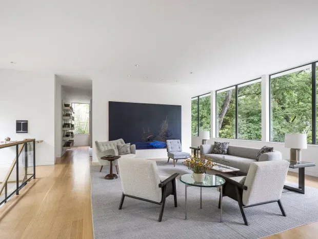 Bright, spacious contemporary living room with floor-to-ceiling windows overlooking leafy trees. The room features pale timber flooring, white walls and ceiling, a large dark media wall, a grey sofa, upholstered lounge chairs, glass coffee tables, and a large neutral rug. An open hallway and shelving are visible at the rear.
