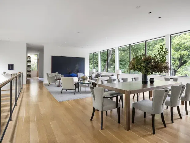 A bright, spacious open-plan living and dining area with full-height windows overlooking leafy greenery. The room features pale timber flooring, a large rectangular timber dining table with upholstered dining chairs, a neutral lounge arrangement, contemporary artwork, recessed ceiling lights, and a dark-framed balustrade along the left side.