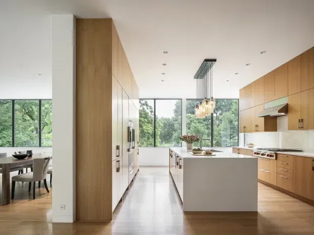 A bright, spacious contemporary kitchen with a large central island, warm timber cabinetry, white stone surfaces, integrated appliances, and a sculptural pendant chandelier. Full-height glazing overlooks leafy greenery, while a dining area is visible to the left. The space has clean linear detailing, recessed ceiling lights, and a warm neutral palette.