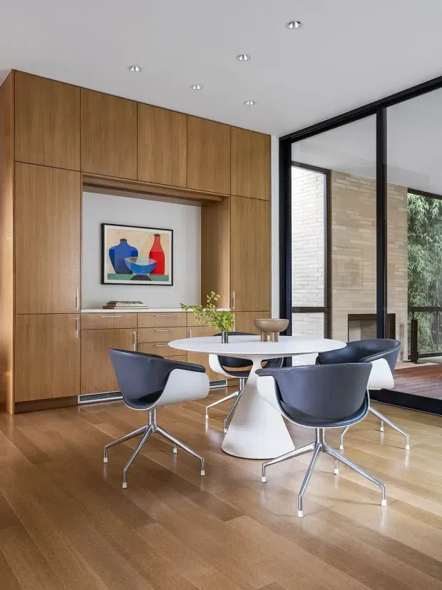 A refined dining area with a round white pedestal table and four sculptural dark-and-white dining chairs. The room features full-height warm timber built-in cabinetry with integrated drawers and display niche, a polished timber floor, white ceiling with recessed downlights, and large black-framed glazed doors opening toward a brick-lined outdoor area. A framed abstract artwork and a small floral arrangement add restrained decorative accents.