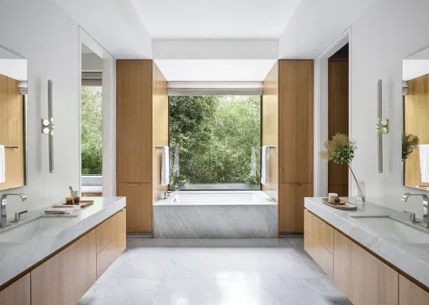 Bright, symmetrical contemporary bathroom with twin floating timber-veneer vanities, white stone benchtops, wall-mounted chrome taps, large mirrors, and tall integrated cabinetry framing a freestanding bathtub. A wide window above the bath overlooks leafy greenery, while pale marble-look surfaces and a skylight-like ceiling opening create an airy, refined atmosphere.