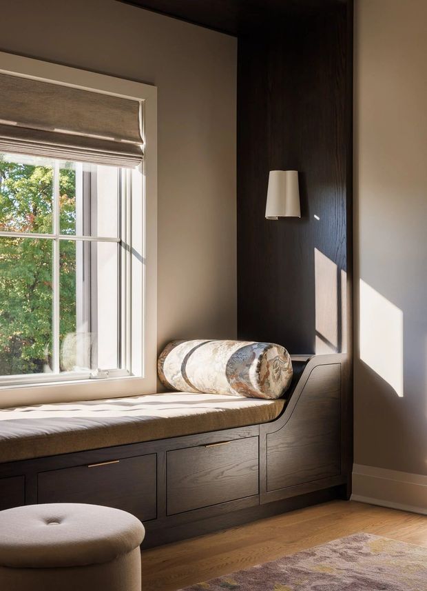 A warm, sunlit built-in window seat with a cushioned bench, dark timber drawer cabinetry, and a vertical dark timber wall panel with a wall sconce. A patterned bolster pillow and upholstered round ottoman add softness, while a large divided window brings in views of trees and strong natural light. The space has a refined, cozy residential interior with custom joinery.