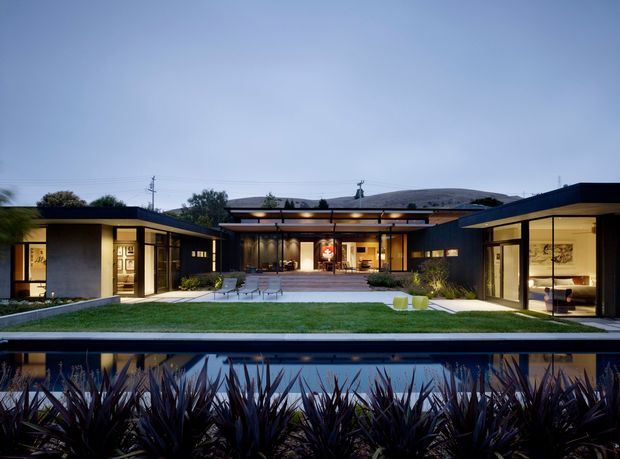 A wide dusk view of a low-slung, single-storey modern residence arranged around a central courtyard and lawn. The dark flat-roofed wings have extensive floor-to-ceiling glazing, warm interior lighting, and a covered central entry or entertaining area. A long reflective pool or water feature occupies the foreground, bordered by dense sculptural planting. The composition emphasizes horizontal lines, indoor-outdoor connection, and a calm contemporary atmosphere.