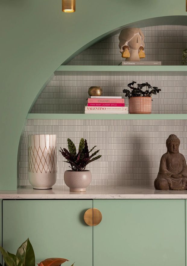 A close-up of a built-in decorative shelving unit framed by a large curved arch. The shelves and lower cabinetry are painted sage green, with a vertically ribbed grey-green tiled or textured wall backing. The display includes books, ceramic vessels, potted foliage, a Buddha sculpture and a sculptural mask, creating a curated, richly layered vignette. A brass-toned circular cabinet pull and woven pendant lights add warm metallic and natural accents.
