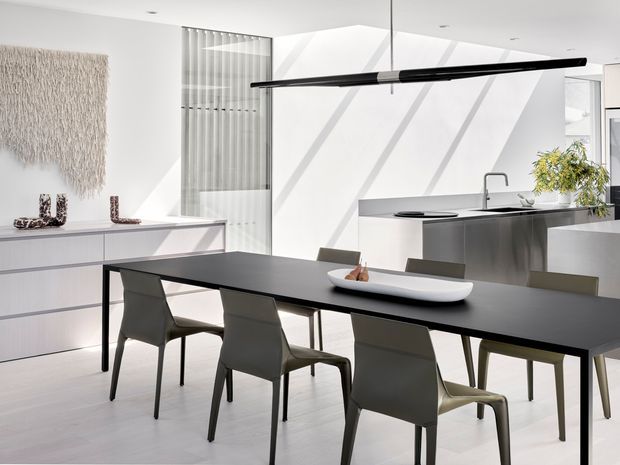 Bright, minimalist open-plan dining and kitchen interior with a long black dining table, sculptural grey dining chairs, pale cabinetry, a dark kitchen island, and a linear black pendant light. White walls and floors are strongly illuminated by sunlight filtered through vertical glazing, creating a clean contemporary atmosphere.