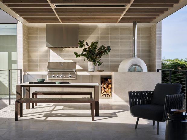 A refined outdoor entertaining area with a built-in stainless-steel barbecue, tiled backsplash, wood-fired pizza oven, integrated firewood storage, and a long timber dining table with benches. A black lounge chair and side table occupy the foreground. The space is covered by a timber-lined slatted ceiling and opens to an outdoor view through a metal balustrade.