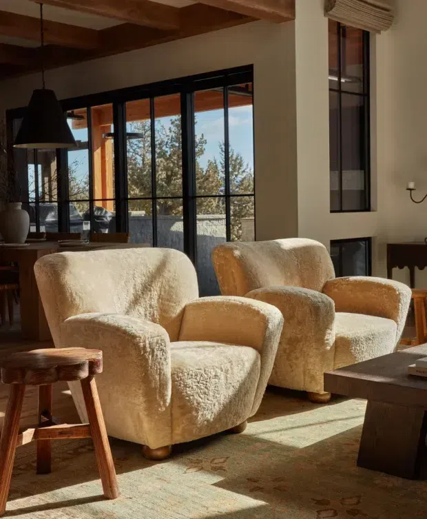Warm, sunlit living room with two cream boucle lounge chairs arranged around a dark coffee table and a rustic wooden side stool. The room features exposed timber ceiling beams, dark-framed floor-to-ceiling glazing, a tall narrow window, a sculptural black pendant light, and earthy timber furnishings. Trees and daylight are visible beyond the glazing.