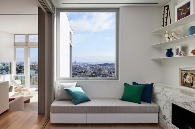 A bright contemporary built-in window seat and lounge nook with a tailored grey upholstered bench, colourful cushions, open white display shelving, and a dramatic marble-clad fireplace surround. Large windows provide expansive city views, while timber flooring and adjacent seating create a connected living area.