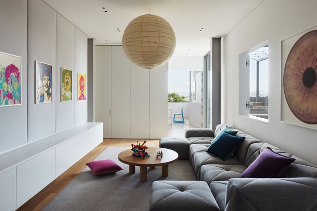 Bright contemporary living room with a large grey sectional sofa, low round timber coffee table, colourful cushions, grey rug, white built-in cabinetry, and a gallery wall of vibrant artwork. A large woven spherical pendant light hangs centrally, while expansive windows and glazed doors provide views and abundant natural light.