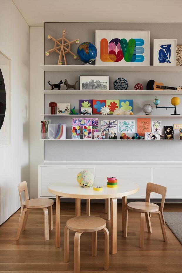 A bright, carefully styled children's dining and activity area with a round light-wood table and matching stools and chairs. A large built-in white display and storage wall fills the background, with open shelves showcasing colourful children's artwork, toys, books, globes and decorative objects. The room has warm timber flooring, white walls and a playful but highly organised aesthetic.