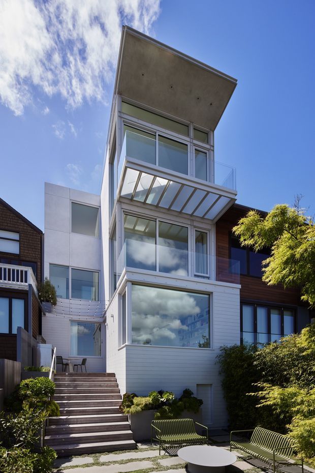 A narrow, multi-storey contemporary residential building viewed from a landscaped courtyard. The facade combines white horizontal cladding, extensive glazed windows and balconies, a projecting concrete roof slab, a translucent canopy, and dark timber cladding on the adjoining volume. Exterior stairs lead to a small paved outdoor seating area with garden planting.