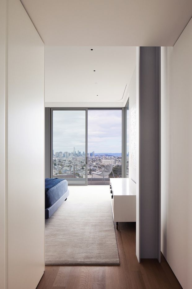 A minimalist, light-filled bedroom viewed from a hallway, with a blue upholstered bed partially visible on the left, a pale rug over warm timber flooring, white built-in cabinetry, and full-height sliding glass doors opening to a balcony with an expansive urban skyline view. The room has a clean contemporary fit-out, recessed ceiling lights, and dark grey window framing.